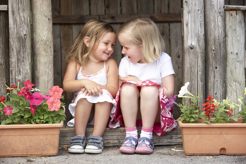 Two little girls sitting on the steps of a wooden shed, sharing a stitched friendship.